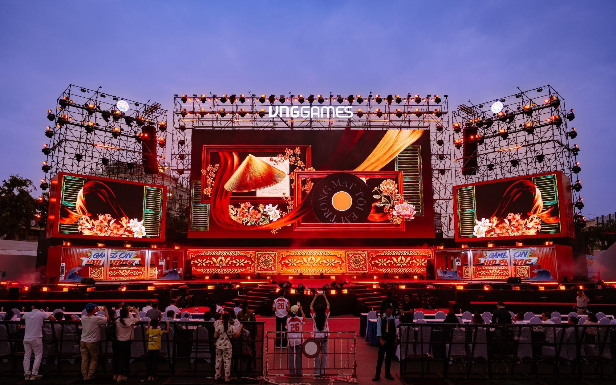 Sân khấu ngoài trời có logo VNGGames, màn hình LED màu đỏ, giàn đèn và người đứng phía trước